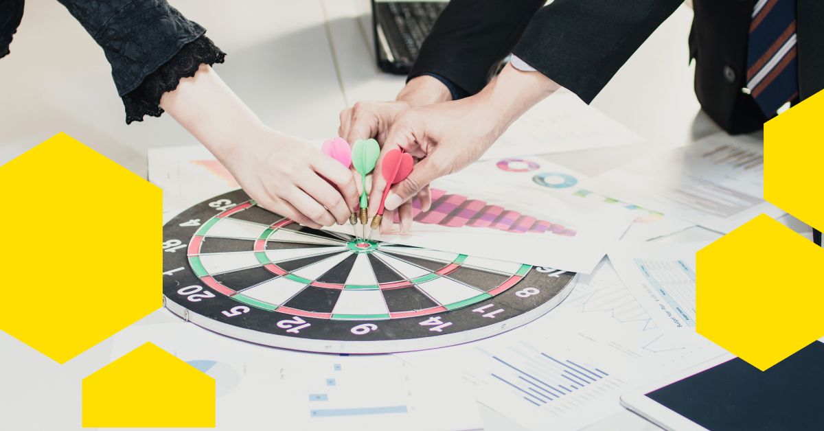 Tři lidé v obchodním oblečení házejí šipky na terč šipkové tabule na stole pokrytém tabulkami a dokumenty, což symbolizuje týmovou práci a společné cíle. Obrázek zdobí žluté geometrické tvary.