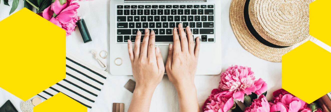 Ruce píšící na notebooku obklopeném květinami a kloboukem, symbolizující kreativní psaní článků.