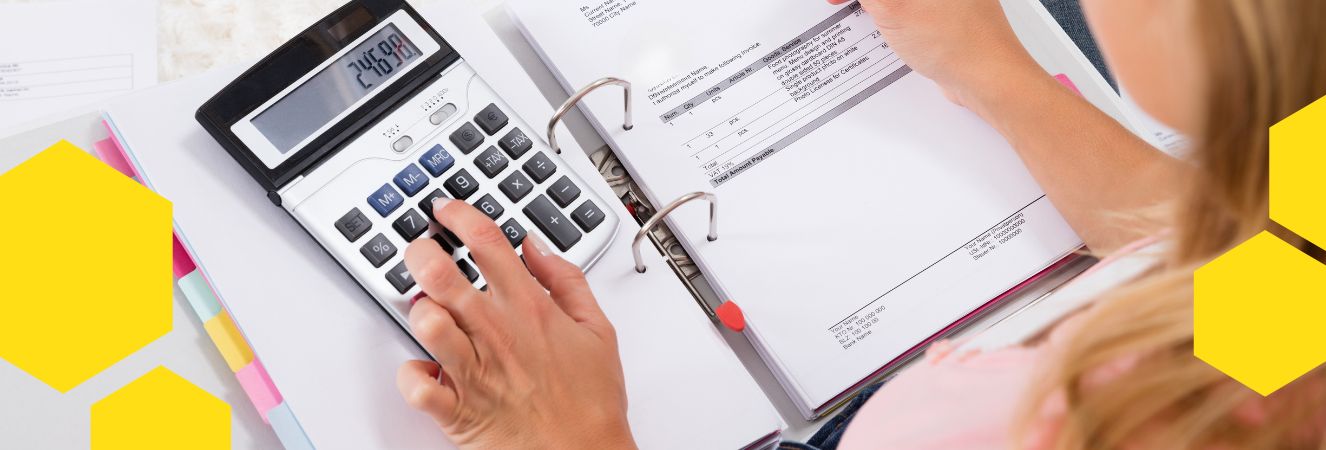 Ruka osoby používající kalkulačku nad otevřenou příručkou s účetními dokumenty. Na stole jsou viditelné pastelové papíry a další kancelářské potřeby.