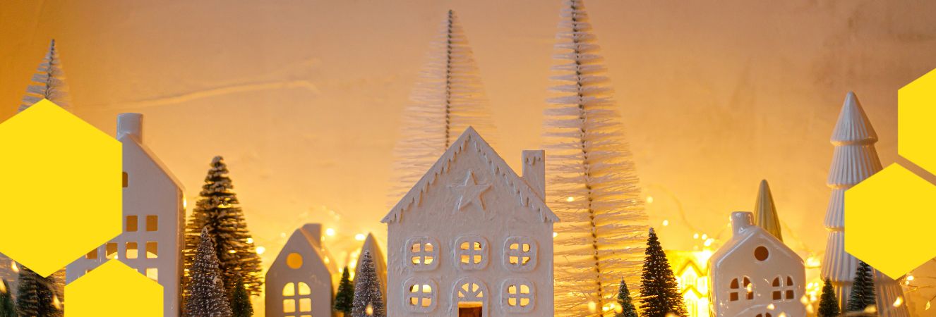 Vánoční dekorace s malými bílými domky, stromečky a vánočními světýlky. Obrázek je rámován žlutými hexagony, které připomínají včelí motiv. Pozadí je jemně rozmazané a vytvořuje teplou vánoční atmosféru.