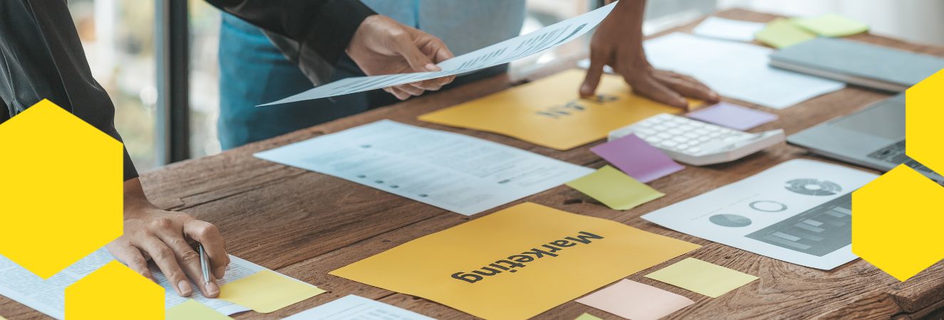 Pracovn&iacute; stůl s dokumenty a pozn&aacute;mkov&yacute;mi l&iacute;stky v různ&yacute;ch barv&aacute;ch, mezi nimiž je pap&iacute;r označen&yacute; n&aacute;pisem &sbquo;marketing&lsquo;. Na stole jsou tak&eacute; tiskoviny a kalkulačka, kter&eacute; se použ&iacute;vaj&iacute; při pl&aacute;nov&aacute;n&iacute; marketingov&eacute; strategie.