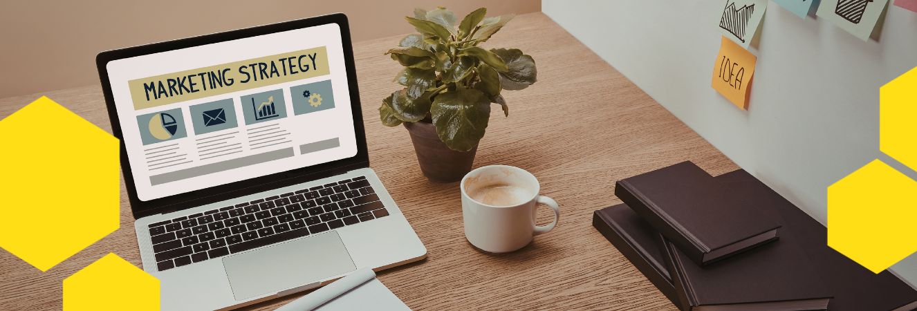 Stůl s notebookem zobrazen&yacute;m na obrazovce s n&aacute;pisem "Marketing Strategy", hrnkem k&aacute;vy, rostlinou v květin&aacute;či a několika z&aacute;pisn&iacute;ky. Na pozad&iacute; jsou barevn&eacute; samolepky s n&aacute;pisy "IDEA".